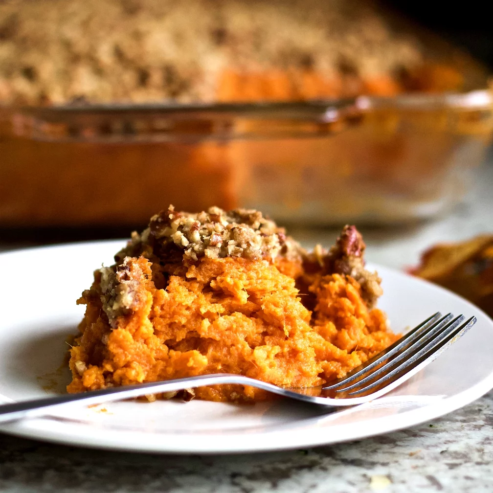Sweet Potato Casserole Serving On A White Plate With The Casserole Dish In The Background.