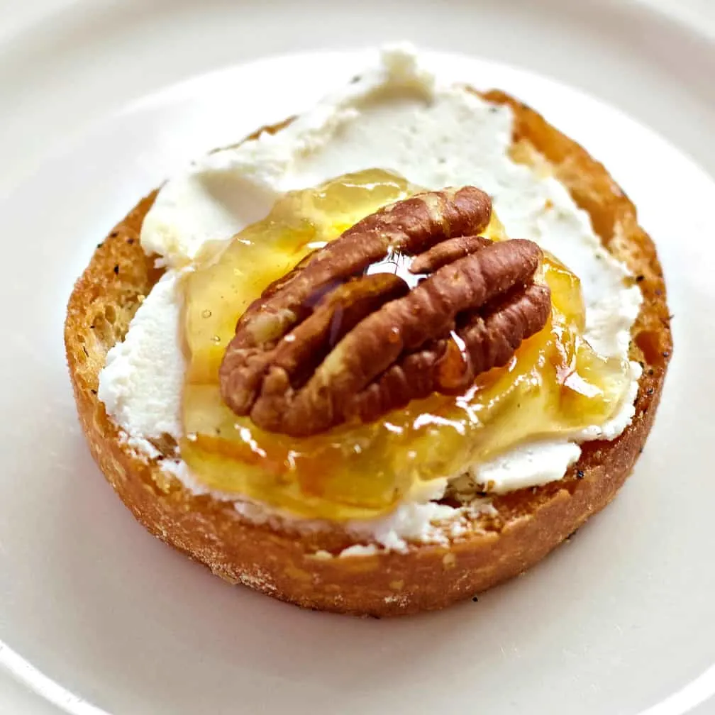 A Single Goat Cheese Crostini Topped With Orange Marmalade And A Whole Pecan. Honey Is Drizzled On Top. Sitting On A White Plate.